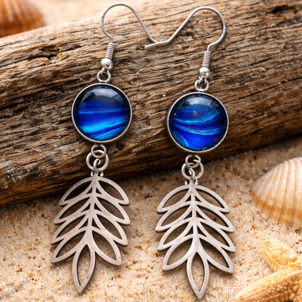 Silver leaf-shaped earrings with blue circular stones on a wooden and sandy background.