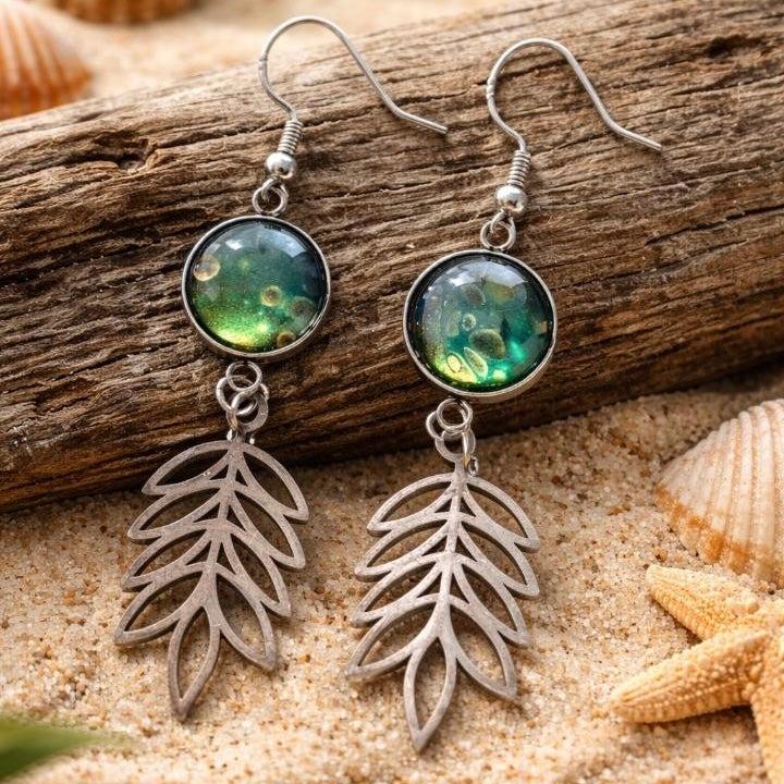 Pair of earrings with leaf design on a sandy background with shells and driftwood.